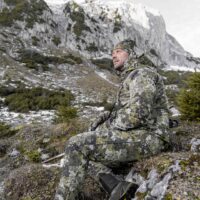 blaser-herren-jacke-huntec-resist-3l.. Die Blaser Herren Jacke Huntec Resist 3L in Camo-Tarrnmuster verfügt über ein 3 lagiges Gewebe und 2 Einschubtaschen sowie 2 Brusttaschen. Die Kapuze ist verstellbare. Auf dem Bild sitzt ein Her auf dem Berg.