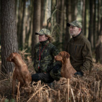 shooterking-herren-softshelljacke-woodlands Die Shooterking Herren Softshelljacke Woodlands ist wendbar mit einer Seite in Grün und der anderen Seite in Camouflage. Sie verfügt über 2 Seitentaschen, 2 Brusttaschen und 1 Armtasche mit Reißverschlüssen. Bild im Wald mit 2 Jägern und 2 Hunden.